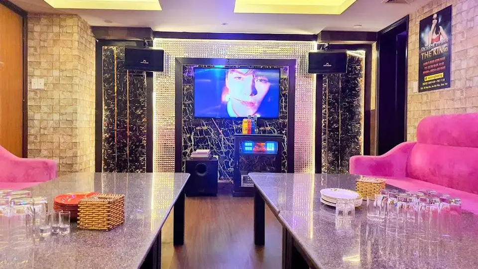 다낭 넘버원 가라오케 그랜드 룸의 대형 LED TV와 블랙 마블 프레임, 핑크 소파 및 완비된 글라스웨어 세팅