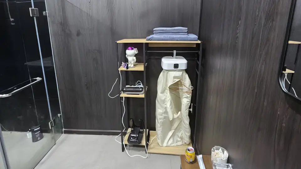 호치민 슈퍼맨 마사지 슈퍼맨 마사지 룸 내 어메니티 코너 — 접힌 타월과 가운이 비치된 오픈 행거 선반, 멀티탭 충전 공간, 소형 프로젝터 및 소품이 정돈된 수납 유닛
